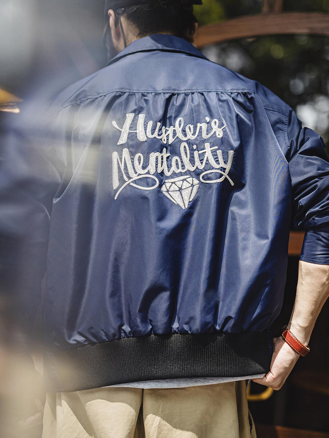 詳細画像
「背面に施された『Hustle is Mentality』スローガンとダイヤモンド型チェーン刺繍のディテール写真。クラシックなデザインと挑戦者の精神を象徴するヴィンテージスタイルのジャケット。」

モデル画像
「モデルがミッドナイトネイビー刺繍ワークジャケットを着用し、防風性のあるコントラストリブ裾やレトロなデザインを活かしたストリートスタイルを披露する写真。」